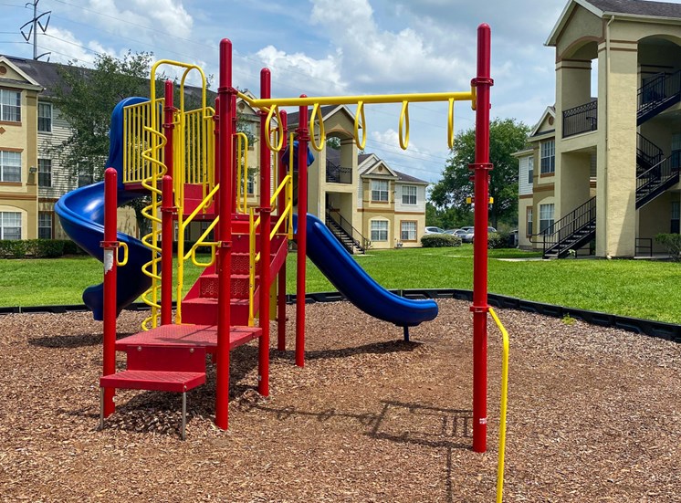 Yellow and Red Playground set with two blue slides in a bed of mulch with buildings and tree in the background